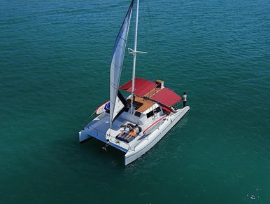 Langsuri catamaran with a red canopy and raised sail, gliding on calm blue waters.
