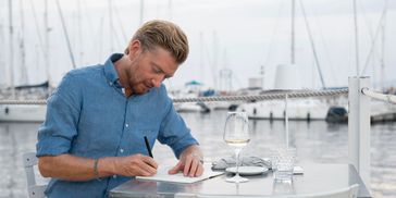 Man writing at a marina-side table with a wine glass, overlooking yachts in the background