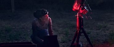 A woman uses a laptop near a red-lit telescope at night outdoors.