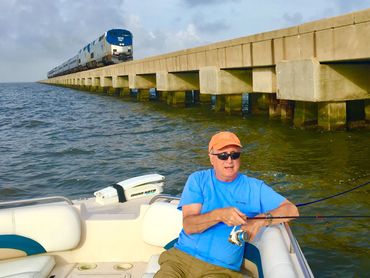 An older man fishes in a boat next to the trestle railroad bridge as an Amtrack train passes by.