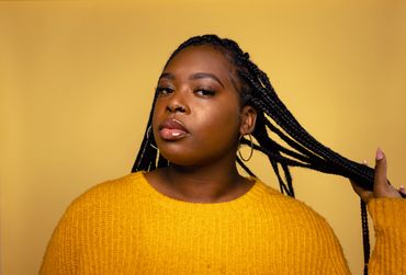 A woman wearing yellow holds her braids in front of a yellow backdrop in a photo studio