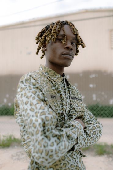 A man wearing neutral color animal print stands with his arms crossed in an abandoned parking lot.