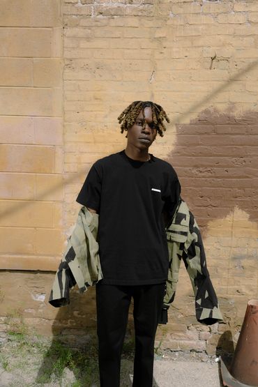A man stands with a patterned jacket falling off of his shoulders in front of a tan brick wall