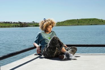A person with a blonde curly afro sits at the edge of a dock over looking water.
