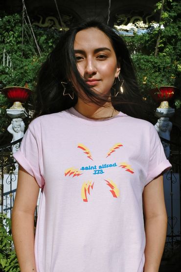 A person wearing large gold star hoop earrings poses with a pink shirt on in front of cherub statues.