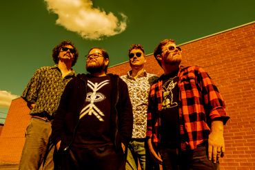 The members of Tiny City stand in front of a brick wall with clouds in the sky above.