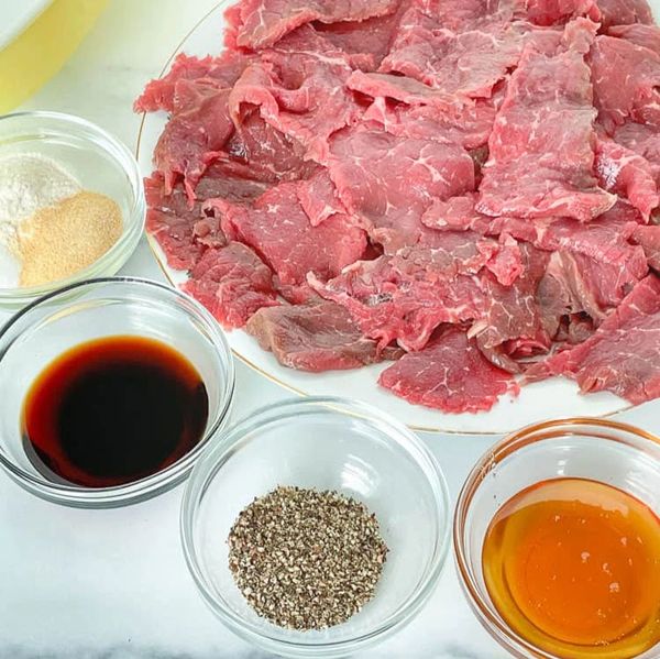 Thinly sliced raw beef with various seasonings in glass bowls on a white surface.