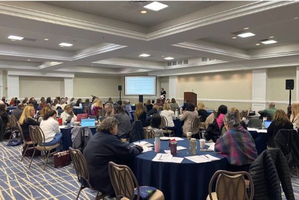 A large group attending a seminar in a conference room with a speaker presenting slides.