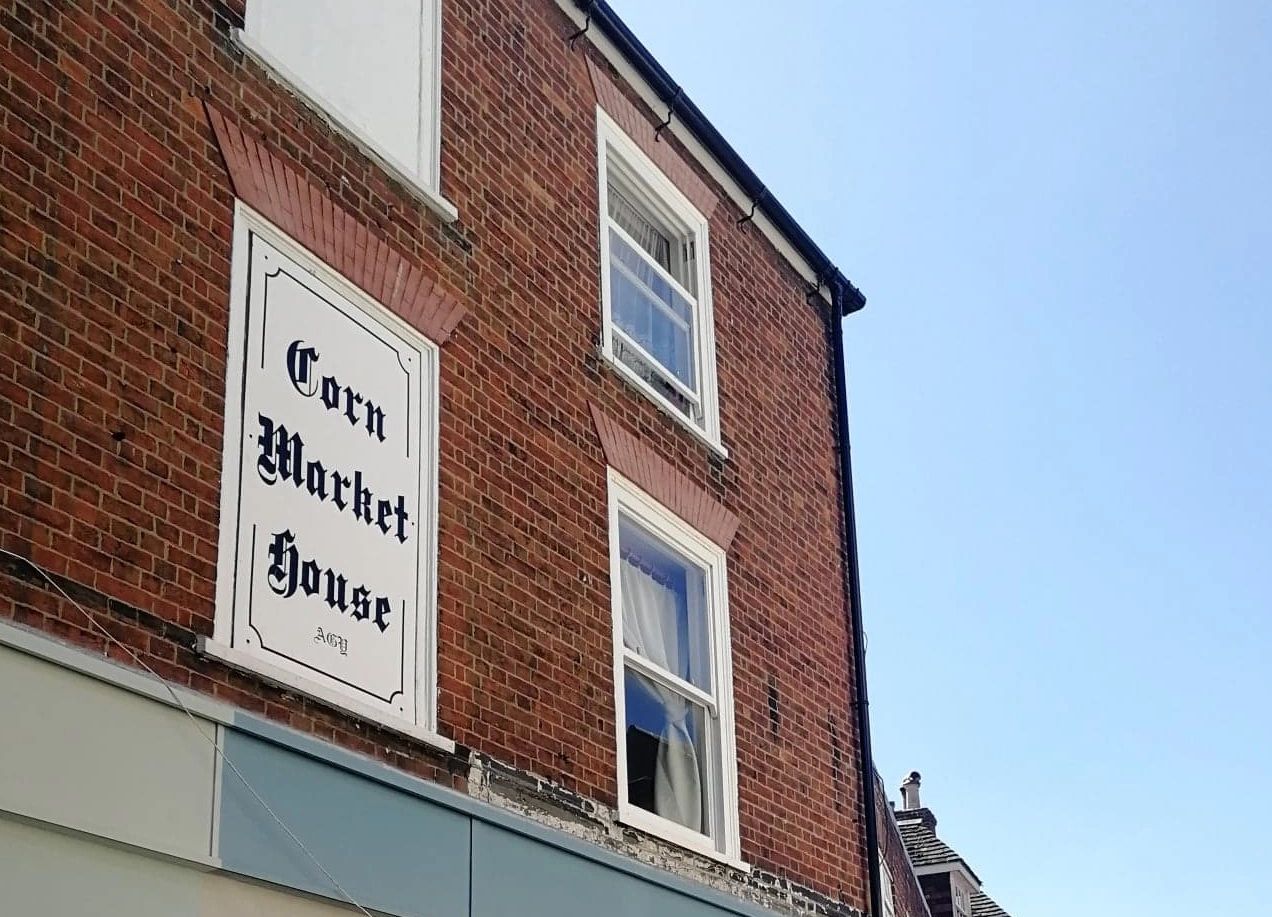 Red brick building against a blue sky with a white sign which has 'Corn Market House' written on it