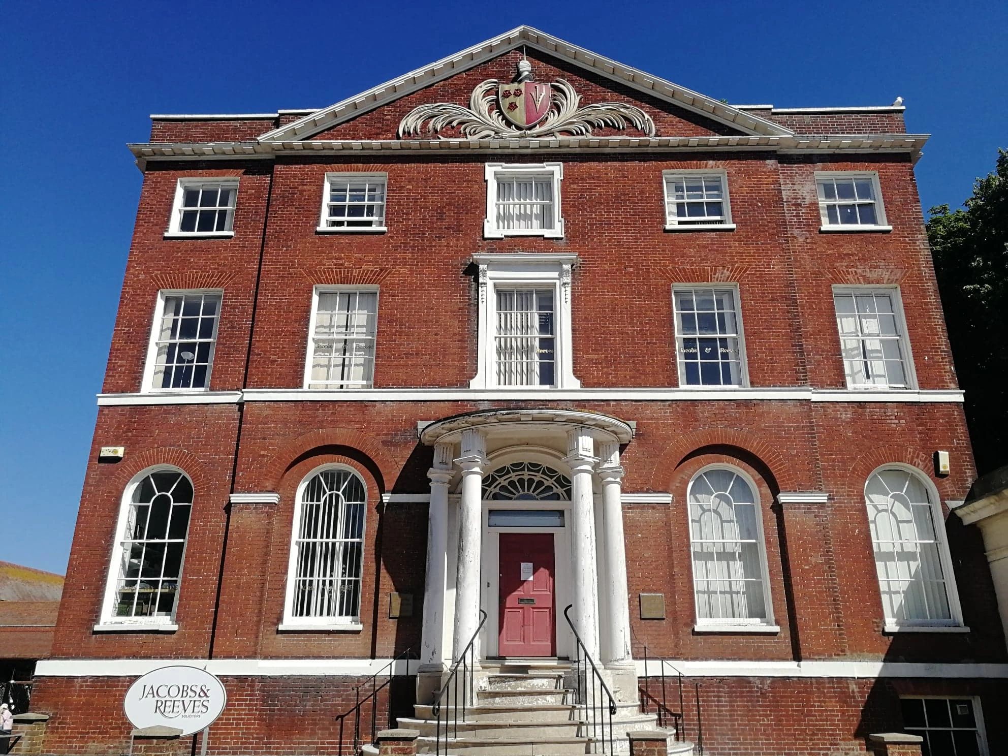 A three story red brick manor house with white pillars around the door and a family crest