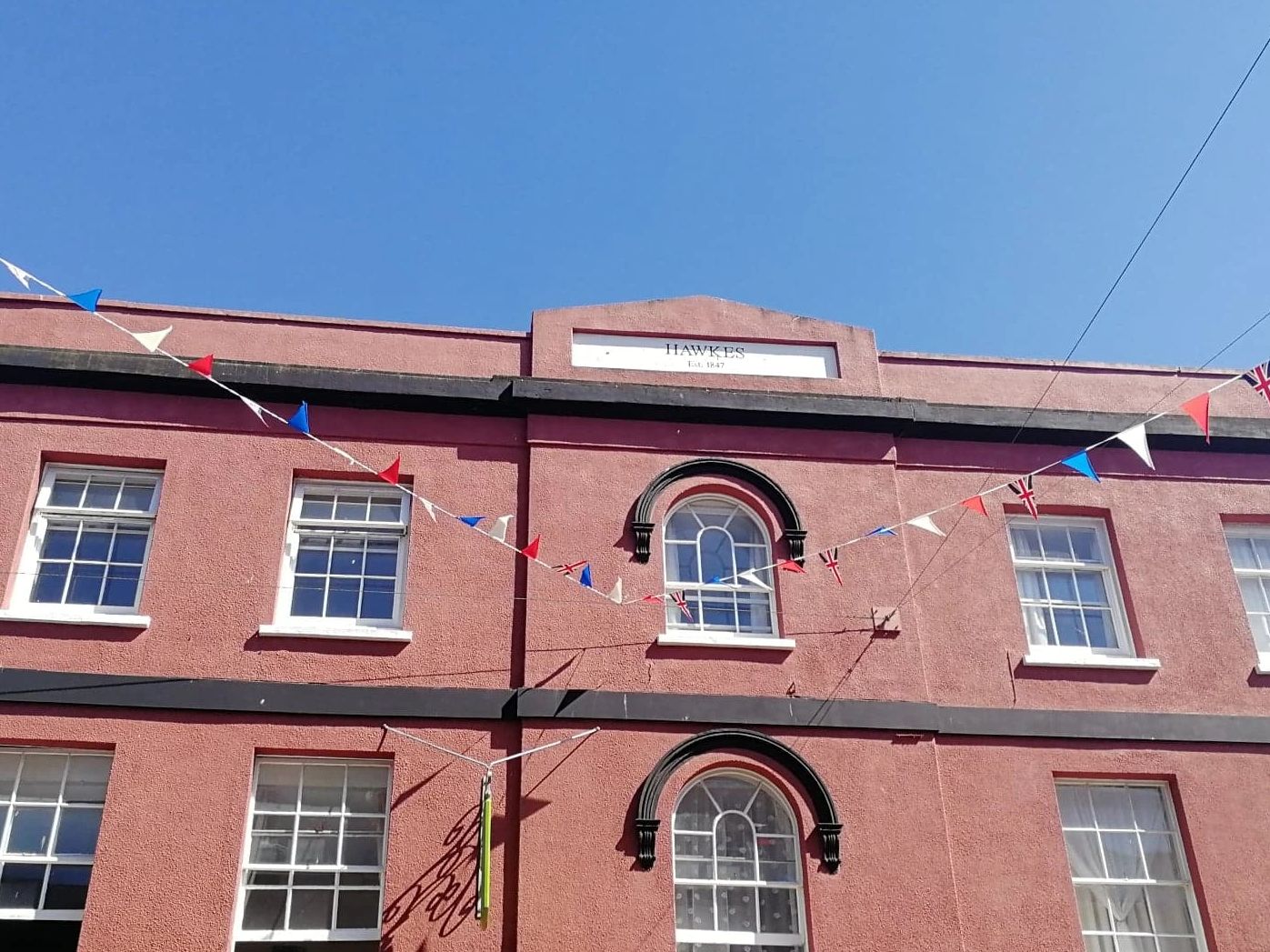 Red painted building with bunting, may windows and a sign saying 'Hawkes ESt 1847'