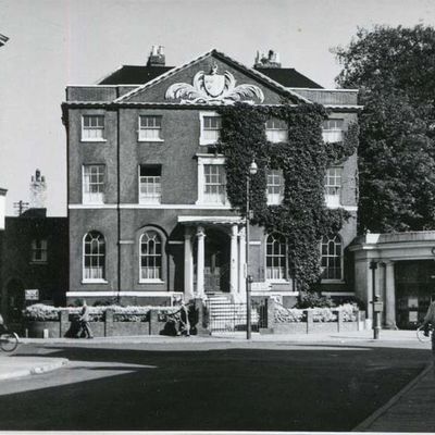 Black & white photo of the brick manor house with people walking on the street in front of the house