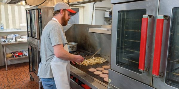 A man cooking burgers and onions on a grill in a commercial kitchen.