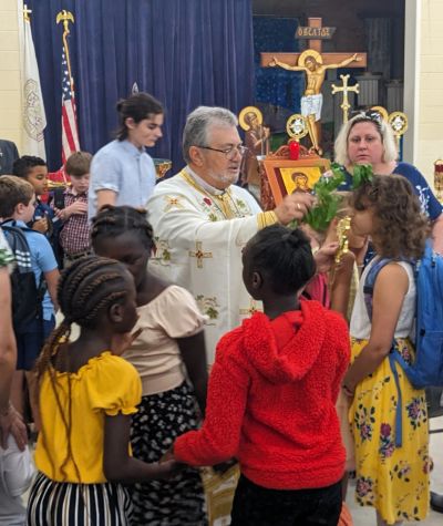 A priest blesses children in a church setting with religious icons in the background.
