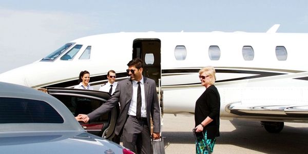 man and woman exiting a car on a tarmac by a private plane and staff