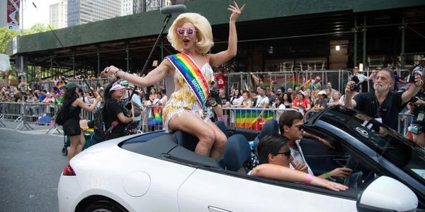 Person in a colorful sash celebrates atop a white convertible during a lively parade.