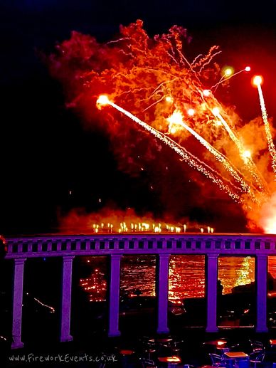 Vibrant fireworks lighting up the night sky above a riverside pavilion.