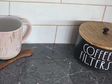 A pink floral mug, wooden spoon, and coffee canister on a kitchen counter.