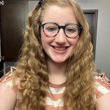 Young woman with long curly hair and glasses smiling indoors.