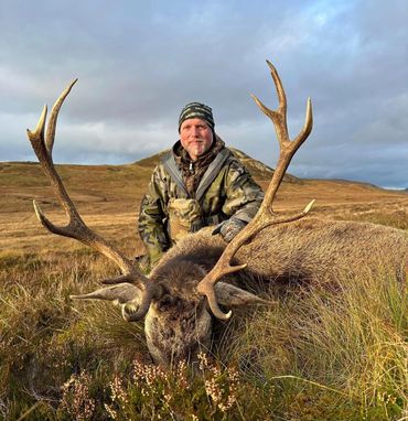Hunter posing with a large stag in a grassy field during cloudy weather.