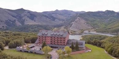 A hotel nestled in lush greenery with mountainous backdrop and a lake nearby.