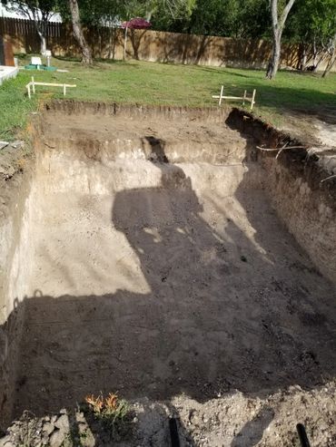 Excavated rectangular pit in a backyard with wooden markers and trees.