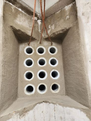 Concrete wall with 12 evenly spaced white circular pipes and hanging copper wires.