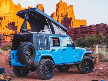 Blue Jeep Wrangler with a pop-up camper top, parked in a campsite.