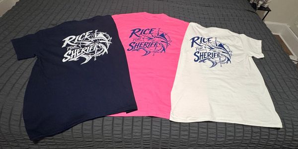 Three "Rice for Sheriff" T-shirts in navy, pink, and white on a bed.