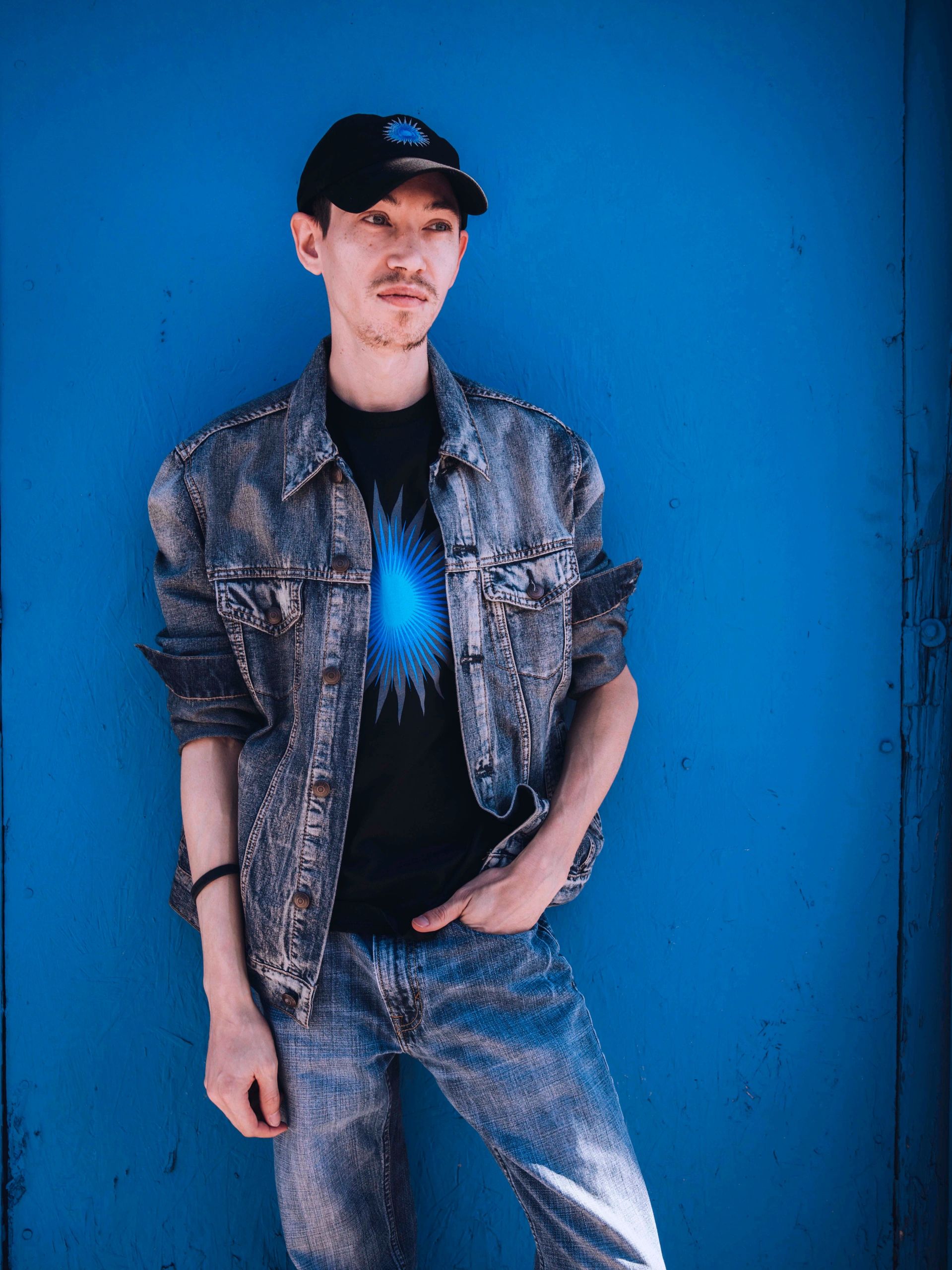 Young man in denim jacket and cap against a blue wall.