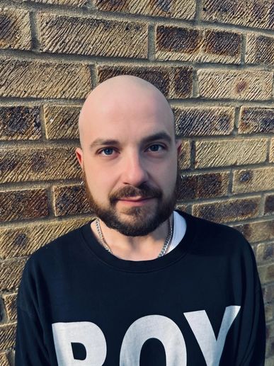 Man with a bald head and beard standing in front of a brick wall.
