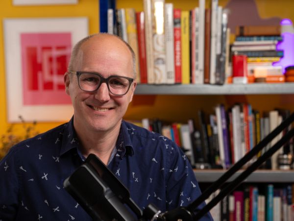 Smiling man with glasses in front of bookshelves and microphone.