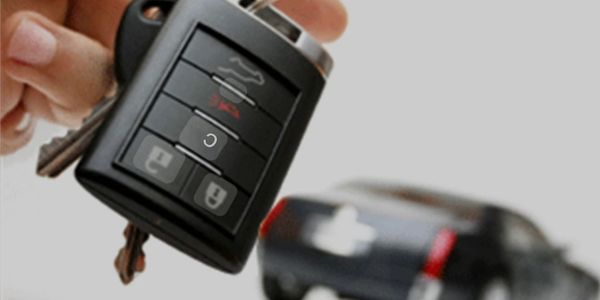 Hand holding car key fob with a blurred black car in background.