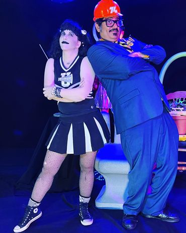 Two people in theatrical costumes posing with arms crossed under blue lighting.