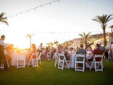 Outdoor dinner event at sunset with guests seated at white tables on grass.