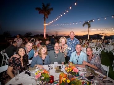 Group of friends smiling at an outdoor evening event with string lights and floral table decorations.
