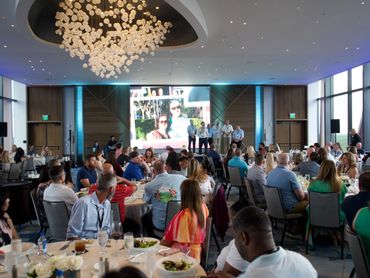 A large group attends a formal indoor event with speakers on stage and a big screen.