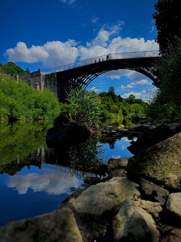 A scenic iron bridge over a reflective river with a bright blue sky and fluffy clouds.
