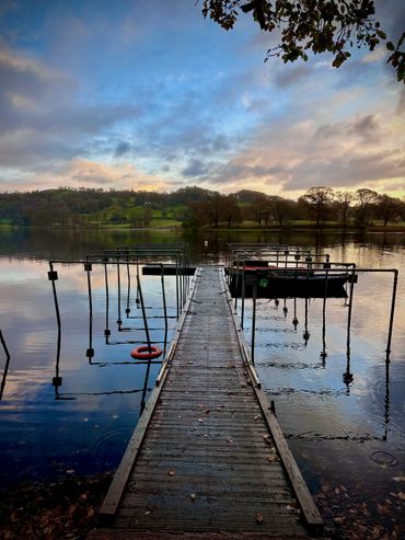 A wooden dock extends over a calm lake at sunset with hills and trees in the background.