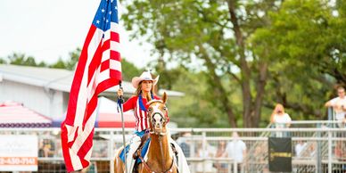 Cowgirl on horseback holding an American flag at an outdoor event.