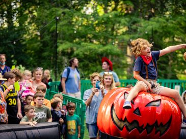 Child joyfully rides a large Halloween pumpkin at a festive event.