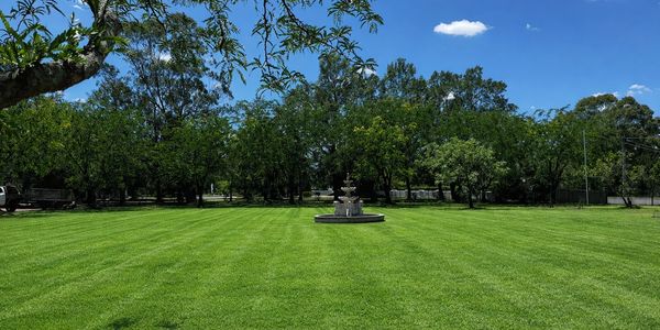 Wide green lawn with a central stone fountain under a clear blue sky.