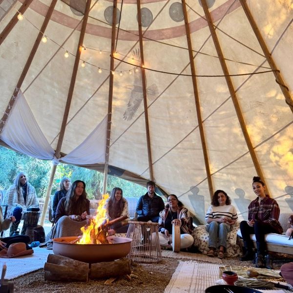 Group of people sitting around a fire inside a tipi, sharing a cozy moment.