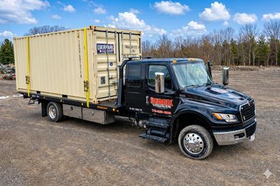 Black flatbed truck carrying a yellow shipping container on a dirt lot.