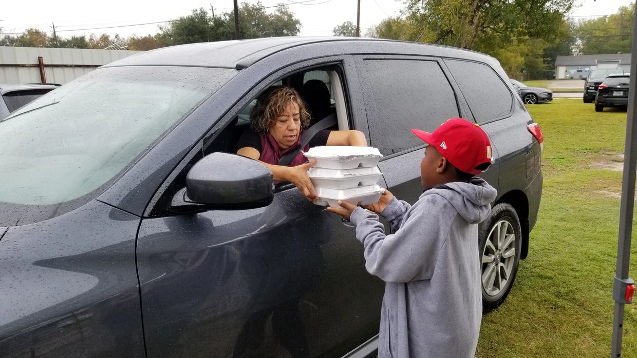 A woman hands food containers to a boy at a car window on a rainy day.