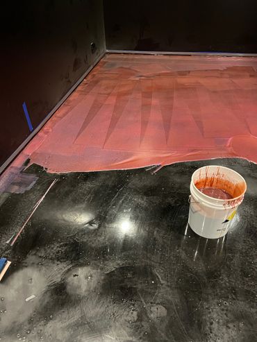 A partially painted floor with a bucket of red paint.