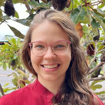 Smiling woman with glasses and long hair in a red shirt, standing outdoors with greenery.