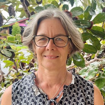 Smiling woman with glasses standing outdoors in front of leafy plants.