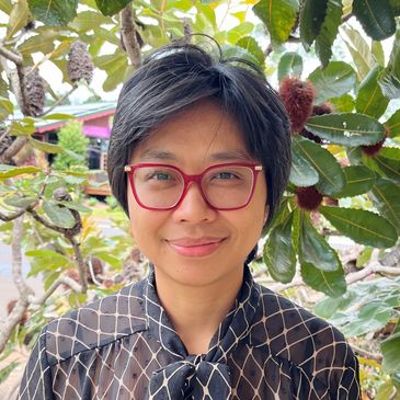 Person with short black hair and red glasses smiling outdoors near leafy trees.
