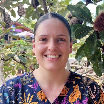Smiling woman in colorful patterned top stands outdoors near leafy plants.